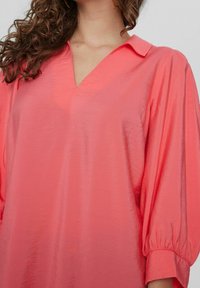 Blusa corallo con scollo a V e colletto, caratterizzata da maniche larghe e gonfiate e un tessuto morbido e testurizzato.