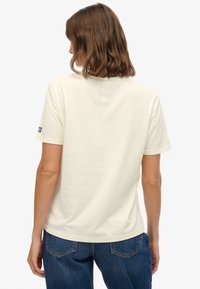 Roomkleurig T-shirt met korte mouwen van zacht katoen, voorzien van een ronde halslijn en een klein logolabel op de linkermouw. Gecombineerd met een donkere spijkerbroek.