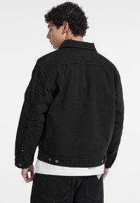 Chaqueta de denim negro con un collar clásico, dos botones en los puños y un acabado texturizado. Usada sobre una camisa blanca, vista desde la espalda.