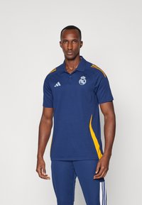 Modrá polo košile s žlutými bočními akcenty, na které je emblém Real Madrid a tři bílé pruhy na levé straně. Krátké rukávy, klasický límec.