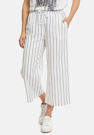 Person iført hvide vidde bukser med sorte lodrette striber, elastisk talje med snøre, afslappede hvide snøresko.