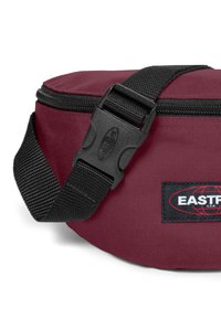 Burgundyröd tygmidjeväska med justerbar svart rem och plastspänne, med ett rektangulärt EASTPAK-logotypmärke.