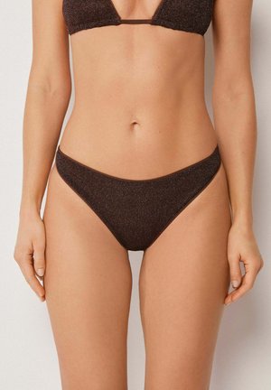 Frau trägt dunkelbraunen glitzernden Bikini-Slip und passendes Oberteil, steht vor einem schlichten hellen Hintergrund, Vorderansicht.