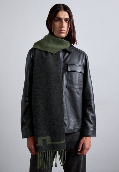 Écharpe en laine de couleur vert foncé et noir, avec un motif géométrique et des bords frangés, portée sur une veste en cuir noir.