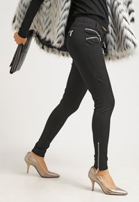 Pantalones negros elásticos con bolsillos con cremallera y cremallera en el tobillo, combinados con zapatos de tacón alto metálicos, la modelo lleva un abrigo de piel sintética.