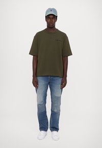 Tricou oversized de culoare verde măslinie, blugi deschiși deteriorați, adidași albi și o șapcă de baseball de culoare albastră deschisă, cu accente brodate.