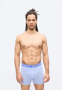 Homme aux longues dreadlocks et silhouette musclée portant un boxer Björn Borg bleu clair, debout devant un fond clair uni.