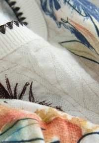 Viacfarebná mäkká tkanina s kvetinovým vzorom v oranžových, modrých a zelených odtieňoch. Má rebrované biele manžety a textúrovaný detail.