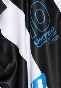 Černobílý úpletový materiál s modrým textem, zobrazující číslo 10, „UNITED“ a „CHAMPIONS AT HEART“ v moderním písmu. Hladký povrch.