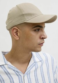 Beige baseballkeps i texturerat tyg, med en styv skärm och justerbar rem på baksidan. Bärs med en randig skjorta.