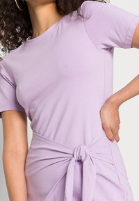 Fioletowa sukienka t-shirtowa z krótkimi rękawami, z wiązanym detalem w pasie i gładką bawełnianą tkaniną. Miękka faktura, casualowy design, subtelny styl.