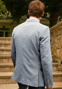 Světle modrý blazer s texturovanou tkaninou, padnoucím střihem a dvěma knoflíky na manžetách. Zadní strana má jeden rozparek. V pozadí schody a kamenná zeď.