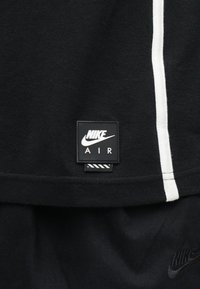 Svart stoff med en teksturert veving; har en hvit stripe og en svart rektangulær etikett med "NIKE AIR" i hvite bokstaver.