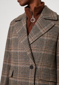 Man die een bruin-grijze geruite wollen jas draagt over een roestkleurige fleece trui met een ronde zilveren ritssluiting.
