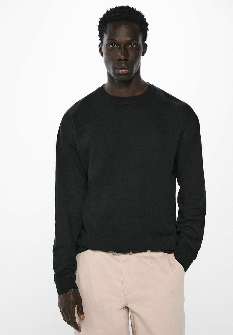 Schwarzes Langarm-Sweatshirt mit Rundhalsausschnitt. Weicher Stoff, gerippte Bündchen und Saum. Kombiniert mit hellen Hosen.