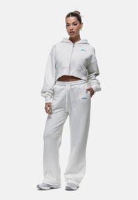 Witte cropped hoodie met rits en geborduurd blauw logo, gecombineerd met bijpassende witte sweatpants met elastische tailleband en witte trekkoorden.