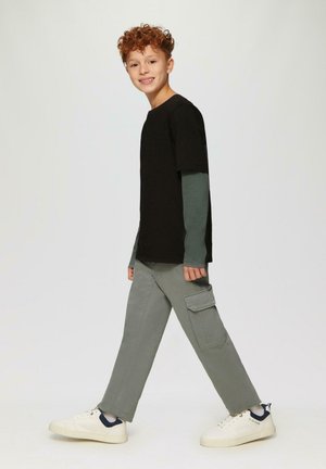 Junge mit lockigem roten Haar, der geht, trägt mehrlagige schwarze und grüne Langarmshirts, graue Cargo-Hosen und weiße Sneakers vor einem einfarbigen Hintergrund.