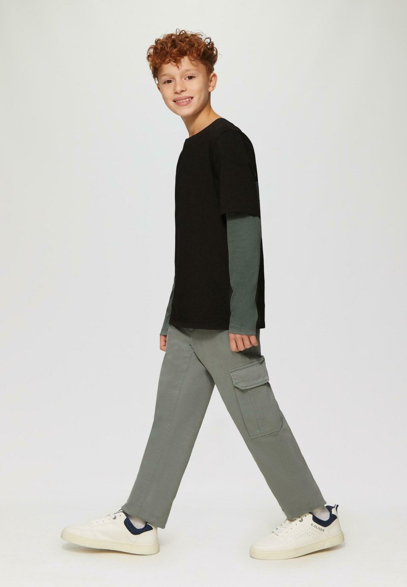 Junge mit lockigem roten Haar, der geht, trägt mehrlagige schwarze und grüne Langarmshirts, graue Cargo-Hosen und weiße Sneakers vor einem einfarbigen Hintergrund.