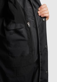 Svart jacka inuti med en dragkedjeficka och tryckt text. Materialet verkar slätt med en matt yta. Snäpplås synligt.