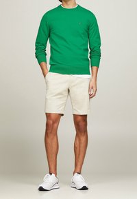 Pull-over vert, manches longues, poignets côtelés ; short beige, ourlet retroussé ; baskets blanches de sport avec accents bleu marine.