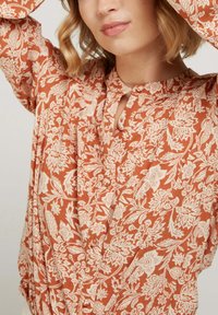 Haut couleur rouille avec un motif floral blanc, présentant un décolleté en u, des manches longues et un tissu texturé qui tombe.