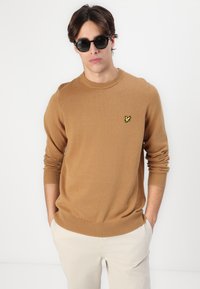Rjav pleten pulover z okroglim ovratnikom, rebrastimi manšetami in robom, vsebuje majhen rumen vezen logotip na prsih. Model nosi črne sončne očala.