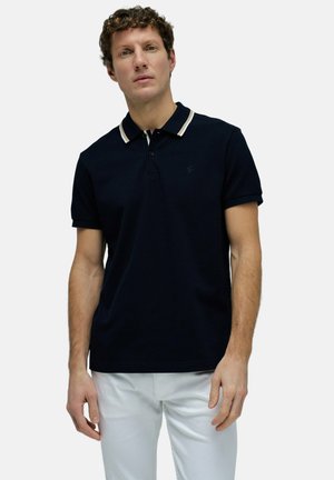 Hombre con camiseta polo negra con ribete blanco en el cuello y pantalones blancos, de pie contra un fondo claro y liso.