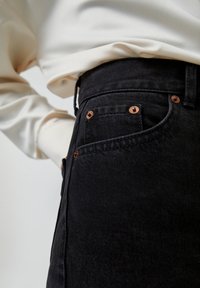Gros plan d'une personne portant un jean en denim noir avec des rivets en cuivre et un haut à manches longues crème rentré.