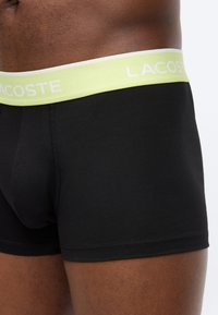 Κοντινή όψη ενός ατόμου που φοράει μαύρα ανδρικά εσώρουχα μποξεράκια με φωτεινά πράσινη ζώνη μέσης με την επωνυμία "Lacoste".