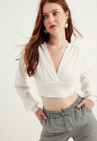 Blusa bianca corta con scollatura a V profonda, tessuto testurizzato e maniche lunghe, abbinata a pantaloni larghi a scacchi in bianco e nero.