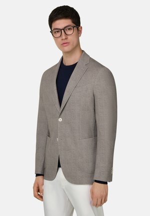Man draagt een zwarte bril, een lichtbruine blazer over een zwart shirt en witte broek, staand tegen een effen lichte achtergrond.