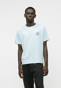 Camiseta de algodón azul claro con cuello redondo, mangas cortas y un pequeño logotipo en marino en el lado izquierdo del pecho. Llevada con jeans oscuros.