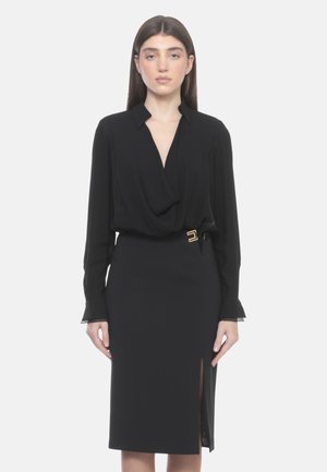 Donna con capelli lunghi e scuri che indossa una camicetta nera a maniche lunghe e una gonna nera al ginocchio con spacco laterale e dettaglio con fibbia dorata.