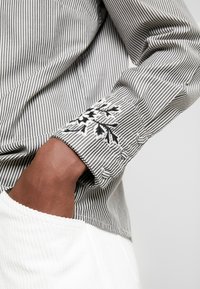 Camicia a righe bianca e nera con maniche lunghe, caratterizzata da dettagli floreali ricamati sui polsini e da un tessuto bianco testurizzato sull'orlo.