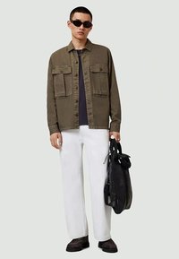 Camisa verde-oliva abotoada com dois bolsos no peito, t-shirt preta por baixo, calças brancas e mochila preta, usada com óculos de sol e sapatos castanhos.