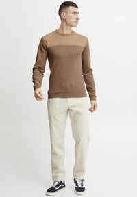 Bruine gebreide trui met een gestructureerd bovenstuk, gecombineerd met crèmekleurige broek en zwarte sneakers. Eenvoudige ronde halslijn en aansluitende manchetten.