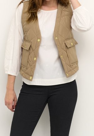 Femme portant un gilet matelassé beige avec des boutons dorés, une blouse blanche à manches longues et un pantalon noir, debout devant un fond uni.