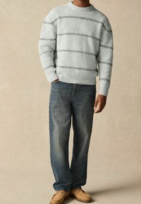 Grijs gestreepte trui met geribbelde textuur, ronde hals; gecombineerd met blauwe spijkerbroek en lichtbruine schoenen. Casual outfit.