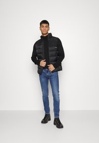 Gilet imbottito nero con colletto alto, tasche con zip, abbinato a un maglione nero e jeans blu, indossato con stivali neri con lacci.