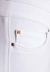 Tejido de mezclilla blanca con un primer plano de un bolsillo delantero, que presenta un pequeño acento dorado y un detalle de hardware plateado. Textura suave visible.