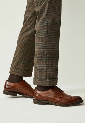 Brogues en cuir marron avec des détails perforés, un bout arrondi et une semelle sombre, associés à un pantalon à motifs carreaux marron.