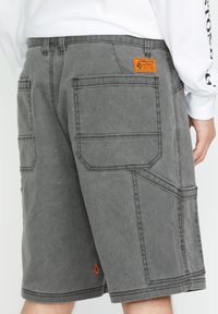 Graue Cargo-Shorts aus strapazierfähigem Stoff, mit mehreren Taschen, einem orangefarbenen Logo-Patch, sichtbaren Nähten und einem lässigen Schnitt.