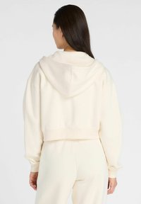 Sweat à capuche coupé fabriqué en tissu beige clair et doux. Présente une grande capuche, des manches longues et un ourlet côtelé. Design simple et sans embellissements.