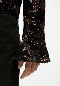 Brown sequined top with a flared cuff and textured black accents; close-up of hand against a dark fabric background.