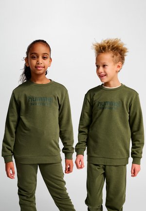 Deux enfants portant des sweat-shirts et pantalons de sport assortis de couleur vert olive de la marque Hummel, debout côte à côte devant un fond uni.