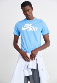 T-shirt de algodão azul claro com grande logótipo branco "JUST DO IT" da Nike, usado com uma camisa branca riscada atada à cintura e calças cinzas.