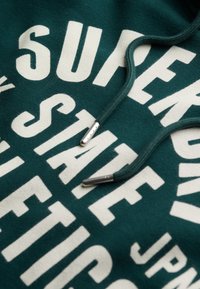 Sudadera de color verde oscuro con gran texto blanco, cordones planos con puntas de metal y una textura suave. Elementos de tipografía en color contrastante y audaz.