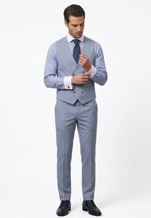 Hombre ajustándose el gemelo, vestido con chaleco y pantalones de traje azul claro, camisa blanca, corbata azul marino y zapatos de vestir negros, sobre un fondo blanco.