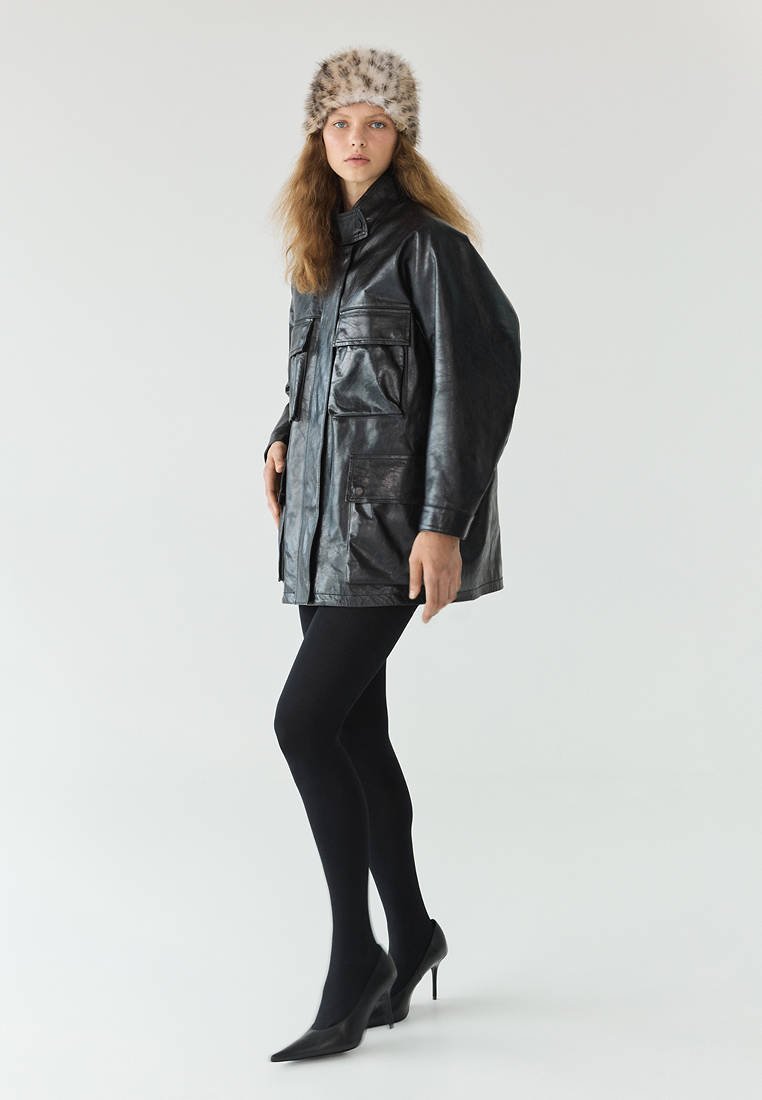Le modèle porte une veste en cuir noire, un collant noir, des escarpins pointus et un bonnet en fourrure, sur un fond de studio uni.