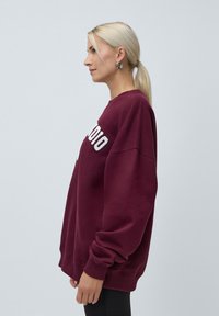 Burgunderfarget oversized genser med rund hals og ribbede mansjetter, med hvit brodert tekst på forsiden. Mykt, teksturert stoff.